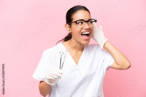Dentist Colombian woman isolated on pink background listening to something by putting hand on the ear