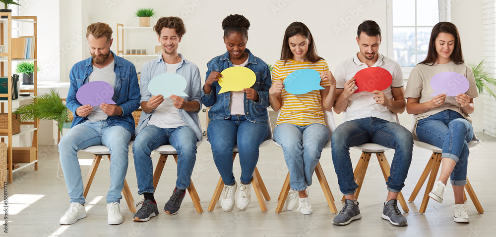 Group of happy diverse mixed race people taking part in public opinion survey, sitting in row in new modern office and looking at colorful mockup text speech balloons they are holding in hands