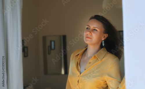 Woman enjoys warm summer breeze while standing in front of the window.