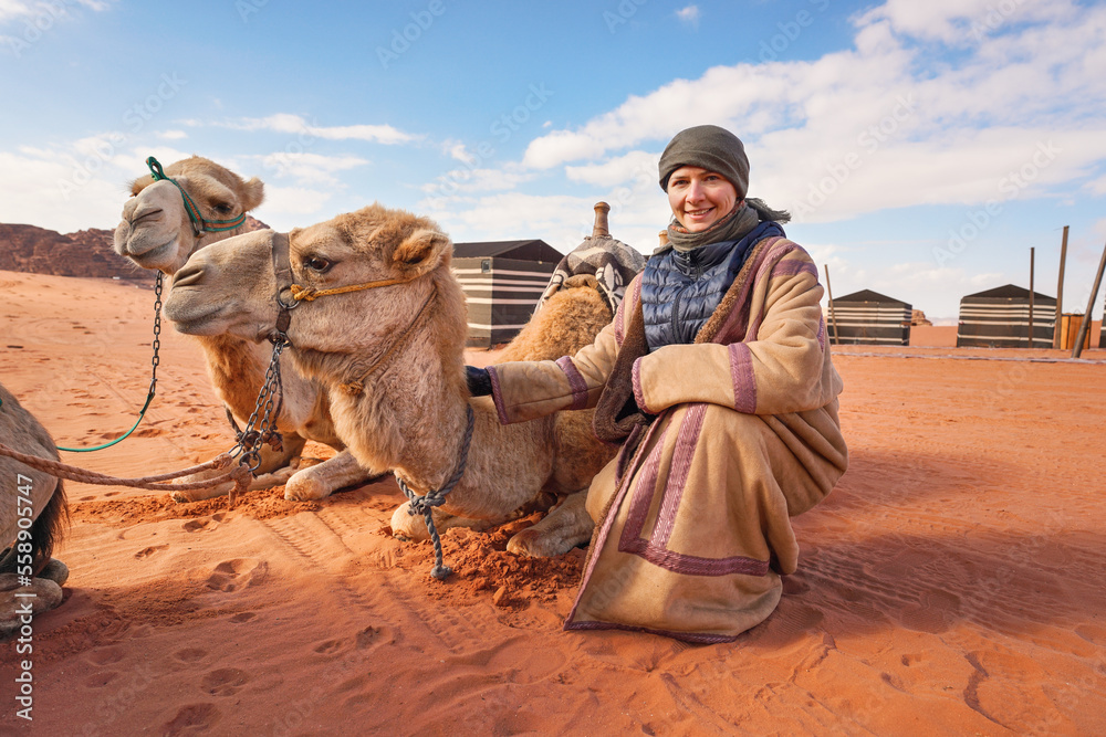 Fototapeta premium Young woman in traditional Bedouin coat - bisht - and headscarf crouching next to two camels laying on red desert ground, smiling, camp tents in background