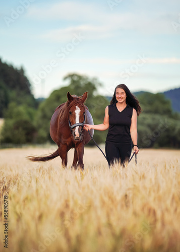 Young woman in black leggings and t shirt walking with brown Arabian horse in wheat field