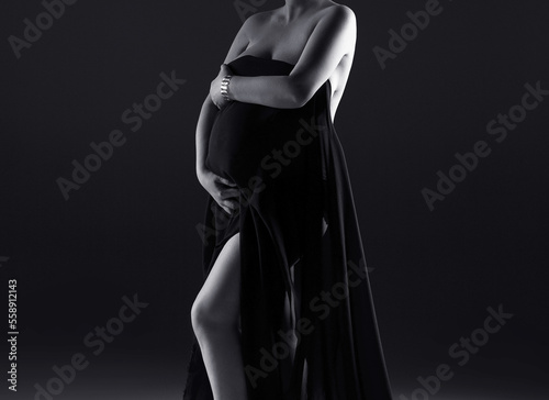 Silhouette of a pregnant woman in a dress on a gray background. A pregnant woman holds her belly. Studio shooting of a pregnant woman. Black and white photo. Woman in black dress.