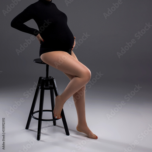 A pregnant woman in a bodysuit is sitting on a chair on a grey background. Studio pregnancy photo shoot. Pregnancy. Shot of a pregnant woman posing in studio. Copy space.