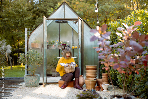Young woman sits with basket full of tulip bulbs for planting in front of tiny glass orangery in garden © rh2010