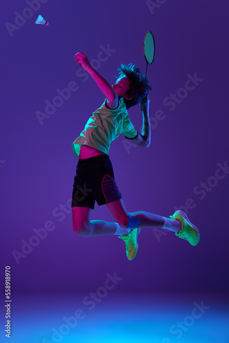 Concentrated teen boy in uniform playing badminton, hitting shuttlecock in a jump over blue purple background in neon ligth