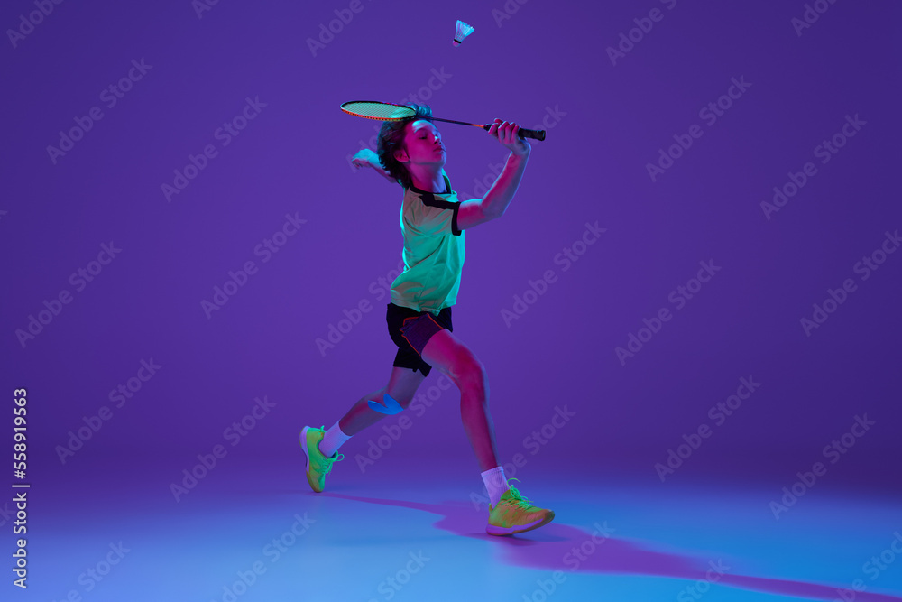 Dynamics. Portrait of teen boy in uniform playing badminton, hitting shuttlecock in a run over blue purple background in neon ligth