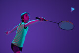 Portrait of teen boy in uniform playing badminton, hitting shuttlecock with racket isolated over blue purple background in neon ligth