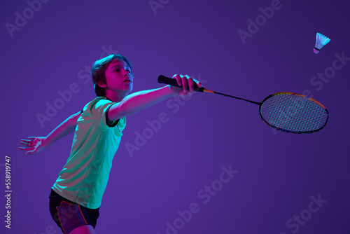 Portrait of teen boy in uniform playing badminton, hitting shuttlecock with racket isolated over blue purple background in neon ligth