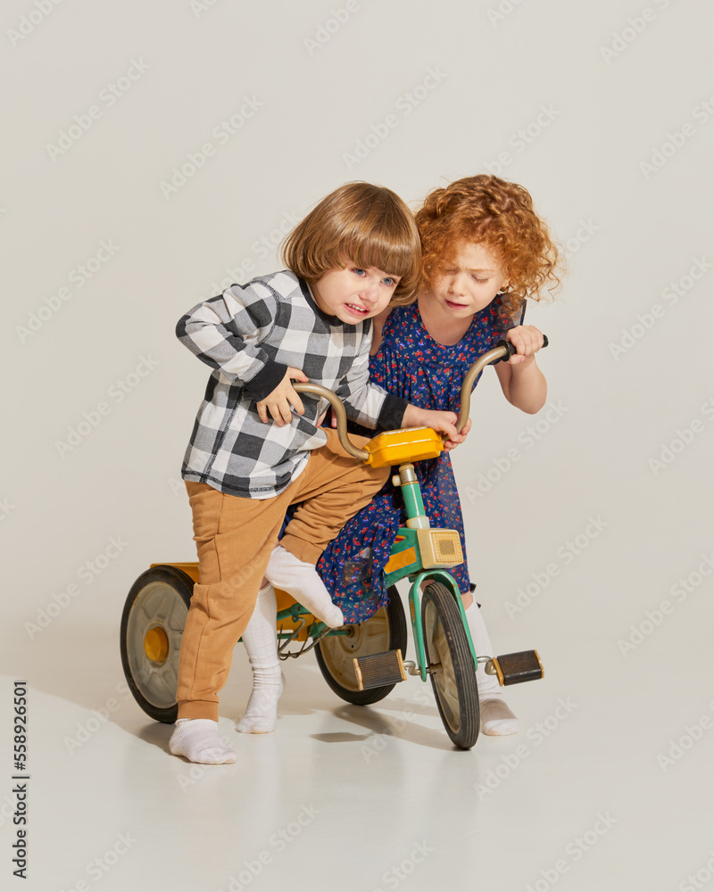 Charming cute kids, habby siblings playing together, riding retro bike isolated over grey background. Concept of carefree childhood, holidays, fun, family, retro style Charming cute kids, habby siblings playing together, riding retro bike isolated over grey background. Concept of carefree childhood, holidays, fun, family, retro style