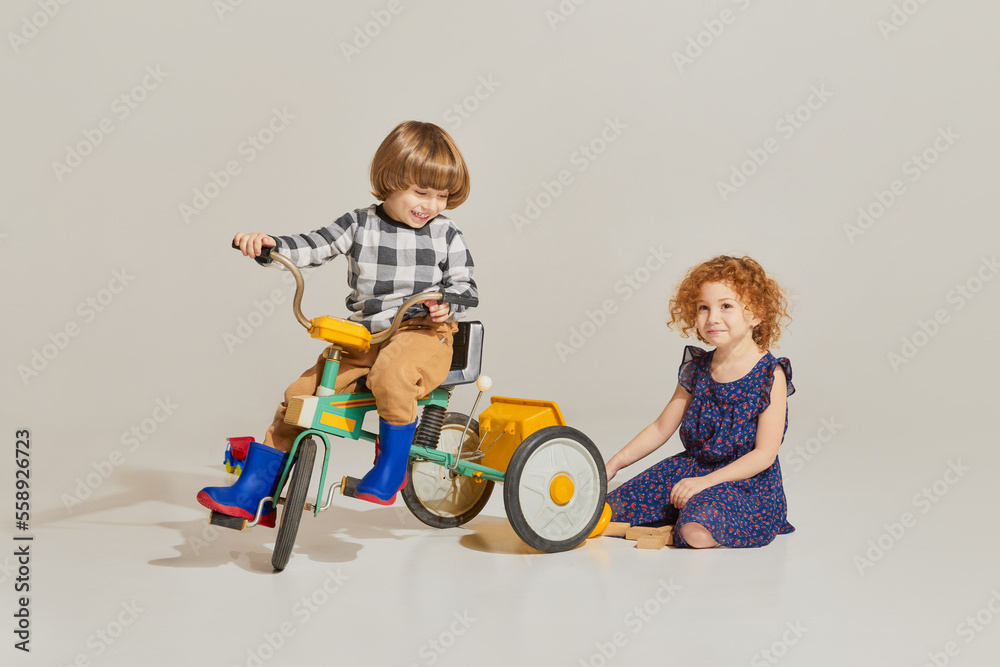 Charming cute kids, habby siblings playing together, riding retro bike isolated over grey background. Concept of carefree childhood, holidays, fun, family, retro style Charming cute kids, habby siblings playing together, riding retro bike isolated over grey background. Concept of carefree childhood, holidays, fun, family, retro style