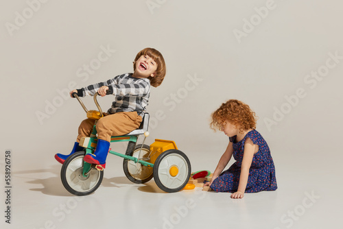 Charming cute kids, habby siblings playing together, riding retro bike isolated over grey background. Concept of carefree childhood, holidays, fun, family, retro style photo