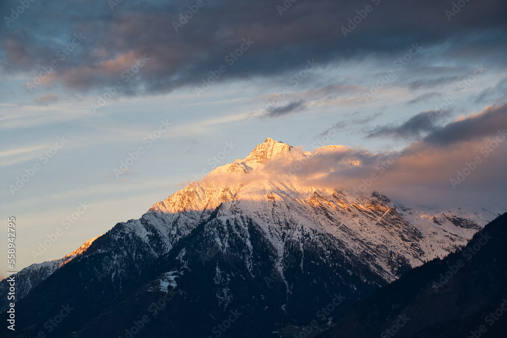 Ein verschneiter Berggipfel im goldenen Morgenlicht mit intensiven, grauen Wolken am Himmel