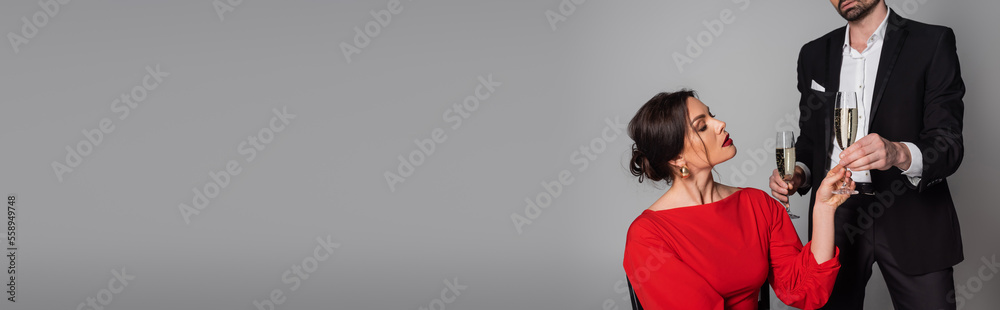 Bearded man in suit giving champagne to brunette girlfriend isolated on grey, banner.