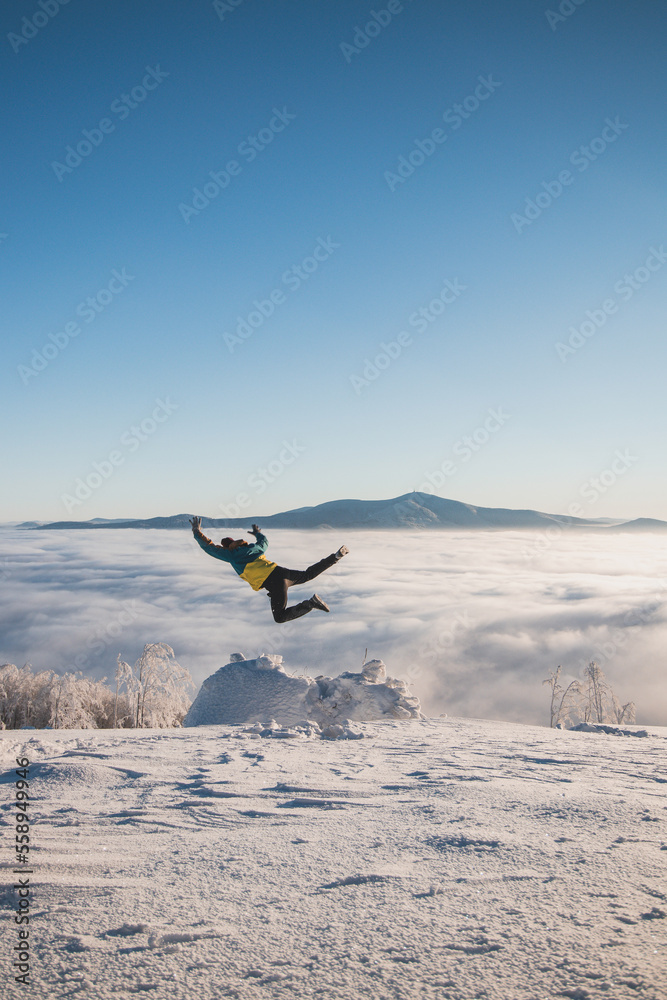Energetic boy in winter clothes jumping from a stump into the fresh snow with a view of the sunrise and clouds rolling along the mountains Energetic boy in winter clothes jumping from a stump into the fresh snow with a view of the sunrise and clouds rolling along the mountains
