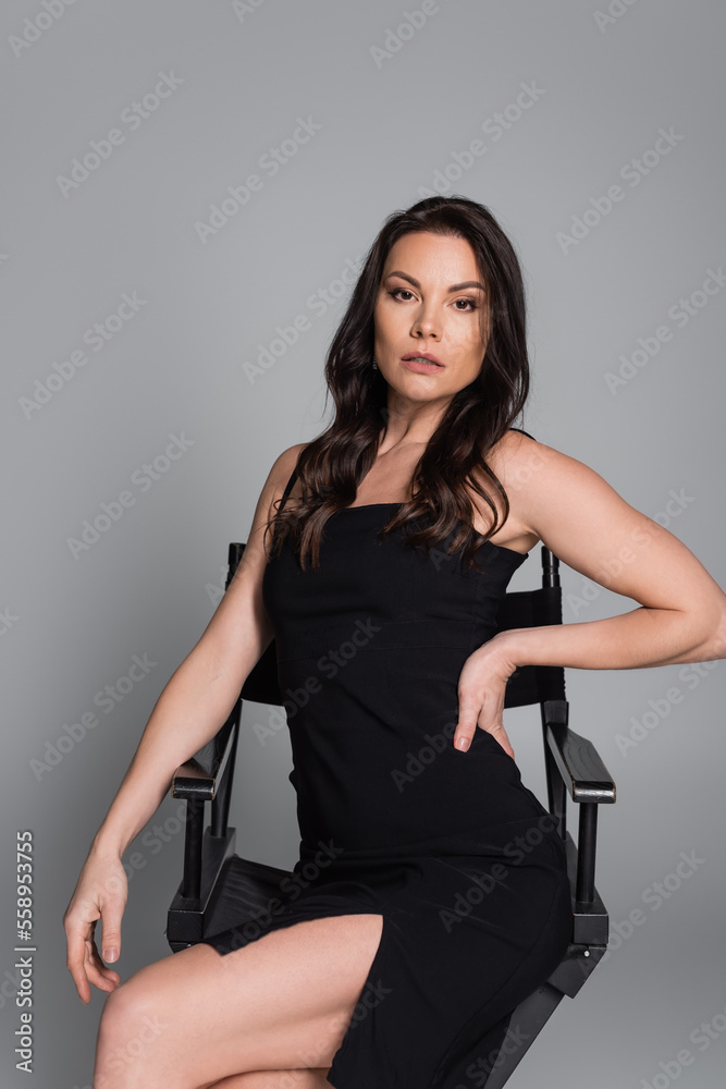 brunette woman in black dress sitting on chair and posing with hand on hip on grey. brunette woman in black dress sitting on chair and posing with hand on hip on grey.