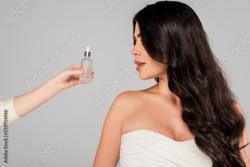 hair stylist holding bottle with oil near woman with shiny hair isolated on grey.