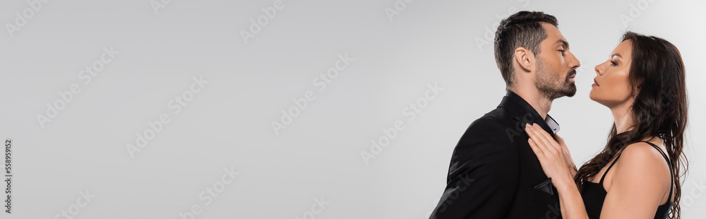 profile of brunette woman in black dress looking at bearded man isolated on grey, banner. profile of brunette woman in black dress looking at bearded man isolated on grey, banner.