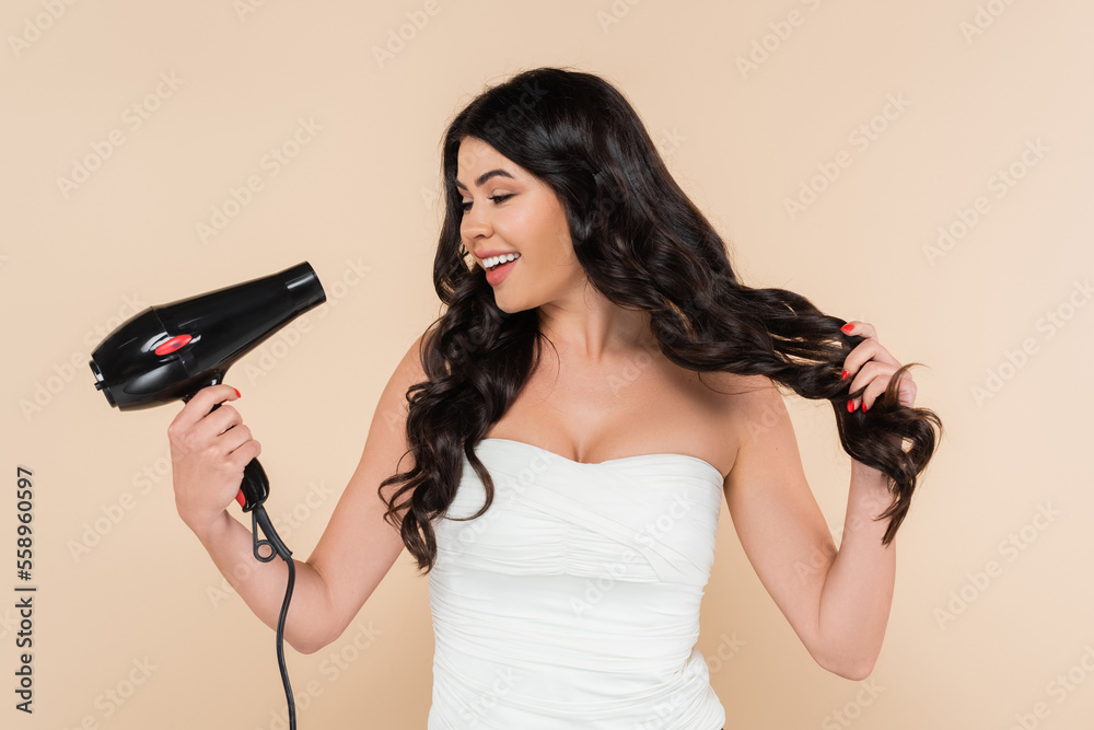 Cheerful brunette woman holding hairdryer and touching hair isolated on beige. Cheerful brunette woman holding hairdryer and touching hair isolated on beige.
