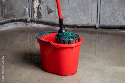 Bucket with mob in flooded basement or electrical room. Cleaning up lots of water on floor from multiple leaks in wall and ceiling. Water damage from rain, snowmelt or pipe burst. Selective focus.