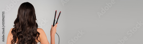 back view of curly woman holding hair straightener isolated on grey, banner.