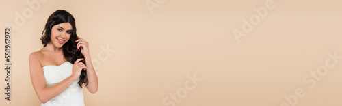 Positive brunette woman in top touching wavy hair isolated on beige, banner.