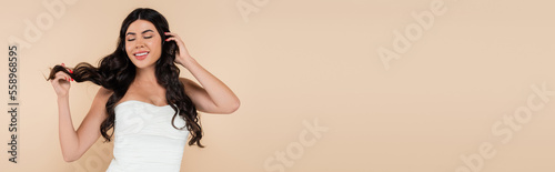 Brunette woman in top smiling while touching hair isolated on beige, banner.