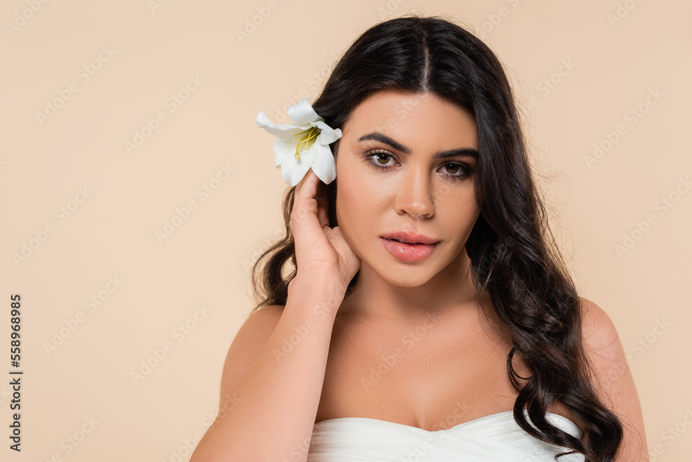 Portrait of brunette woman with lily flower in hair looking at camera isolated on beige.