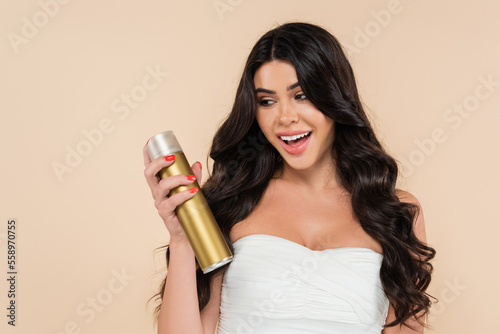 Excited woman with wavy hairstyle holding hairspray isolated on beige.