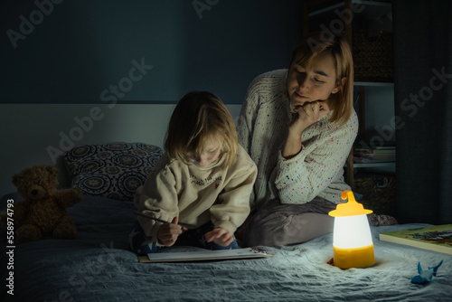 Mother and little daughter studying and drawing in a complete darkness during electricity outage. Little girl uses camping lantern to do her homework during blackout. Energy crisis concept