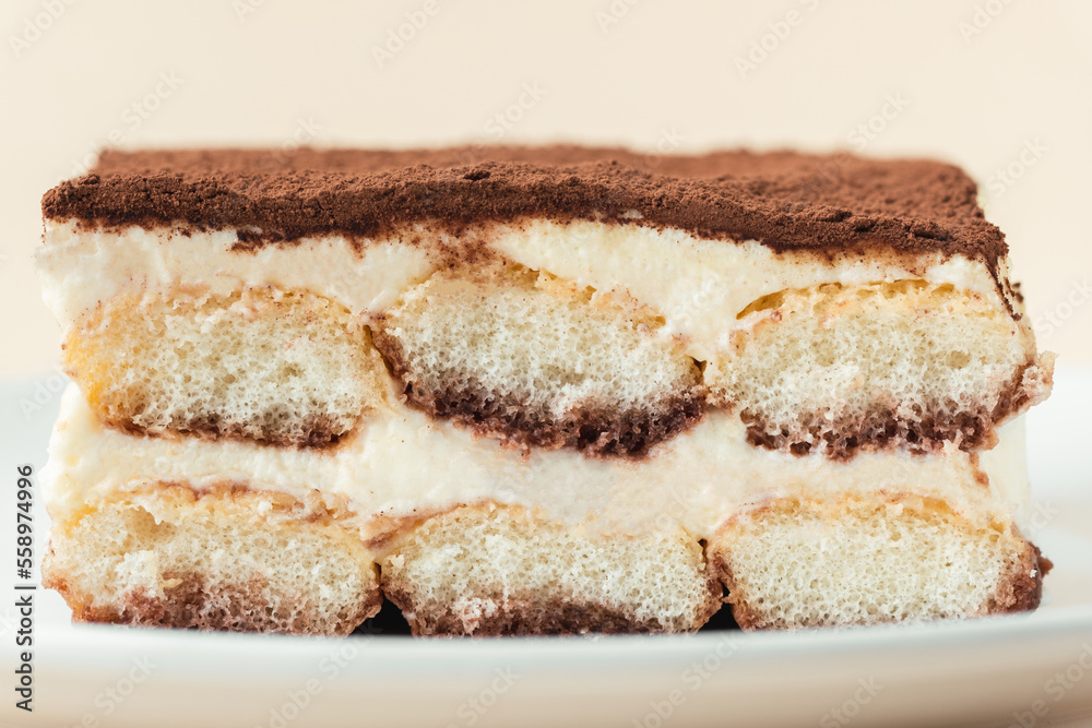 Homemade tiramisu cake on white plate against beige background. Traditional Italian no-bake dessert made of savoiardi, filled with mascarpone cheese, coffee espresso and sprinkled with cocoa powder
