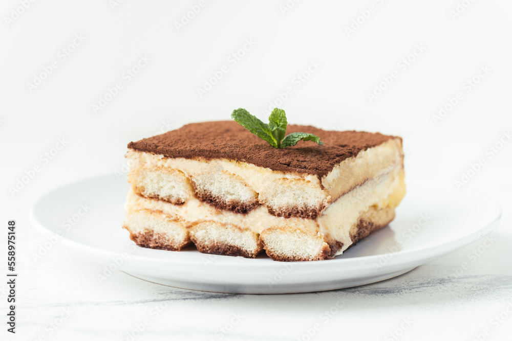 Homemade tiramisu cake on white plate against white background. Traditional Italian no-bake dessert made of savoiardi, filled with mascarpone cheese, coffee espresso and sprinkled with cocoa powder Homemade tiramisu cake on white plate against white background. Traditional Italian no-bake dessert made of savoiardi, filled with mascarpone cheese, coffee espresso and sprinkled with cocoa powder