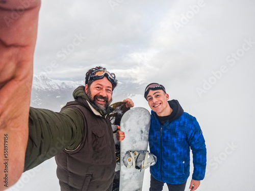 Father and son in friendship enjoying together winter holiday vacation doing snowboard and taking selfie picture with phone. Young and mature smiling and having fun in outdoor mountains leisure