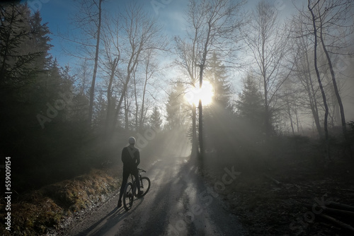 Fotografia Person resting on a mountain bike ride uphill reaching sun piercing through winter fog or cloudy weather