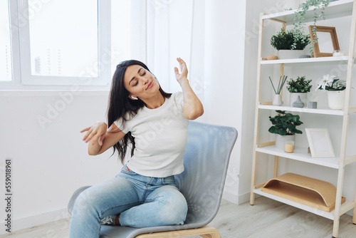 Woman sitting in a chair listening to music and dancing with wireless headphones at home in jeans and a white T-shirt, fall lifestyle comfort