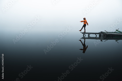 Side view of fashioned young woman sitting on wooden dock looking at view on a misty morning. Female hipster relaxes on the edge of jetty admiring foggy landscape. Wonderful, playful nature getaway