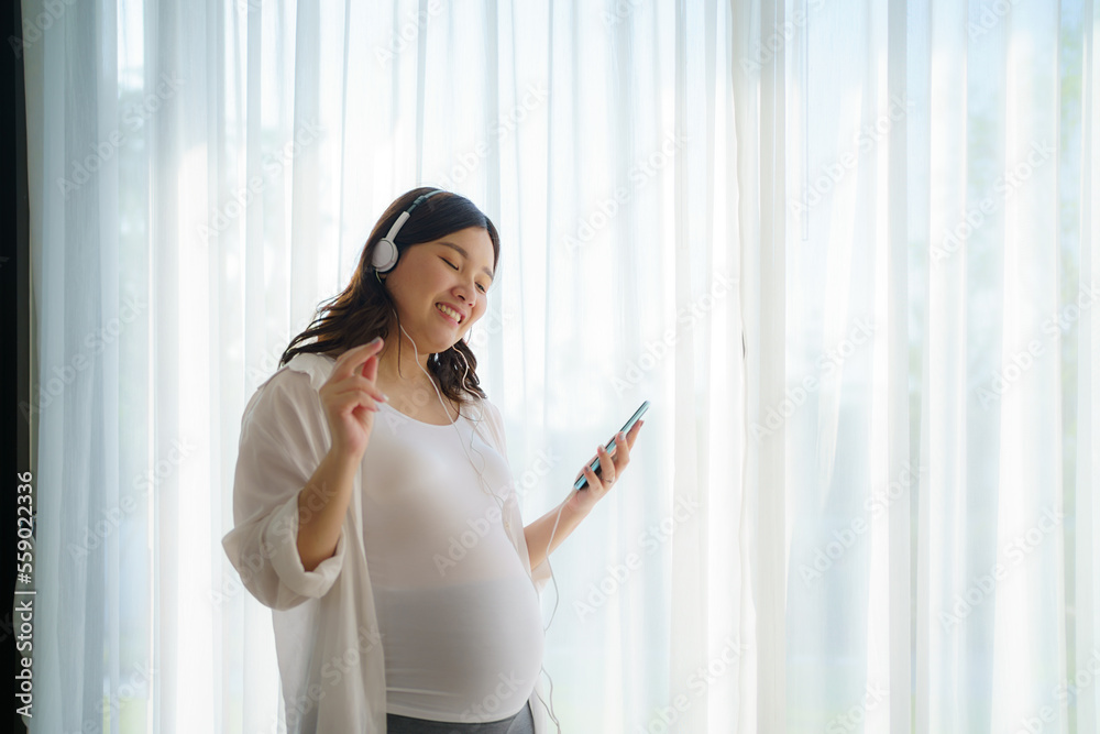 Portrait of pregnant Happy Asian woman standing in living room at home and using smart phone for listening music in headphones. Portrait of pregnant Happy Asian woman standing in living room at home and using smart phone for listening music in headphones.