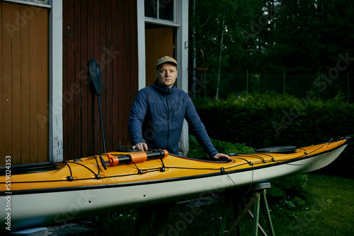 nordic finnish caucasian white outdoor man taking care of his kayak that is yellow wearing blue in front of an old vintage barn during summer evening wearing a cap in finland