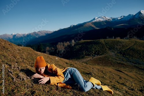 Woman lying on a hill on the grass rest smile with teeth looking at the mountains in the snow in spring in a yellow raincoat and jeans happy sunset trip on a hike, freedom lifestyle 