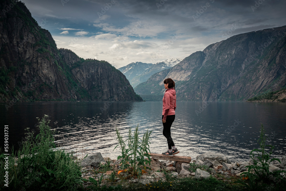Woman standing at waterfront of lake in the mountain landscape Eidfjord in Norway, looking into the fjord, clouds in the sky
