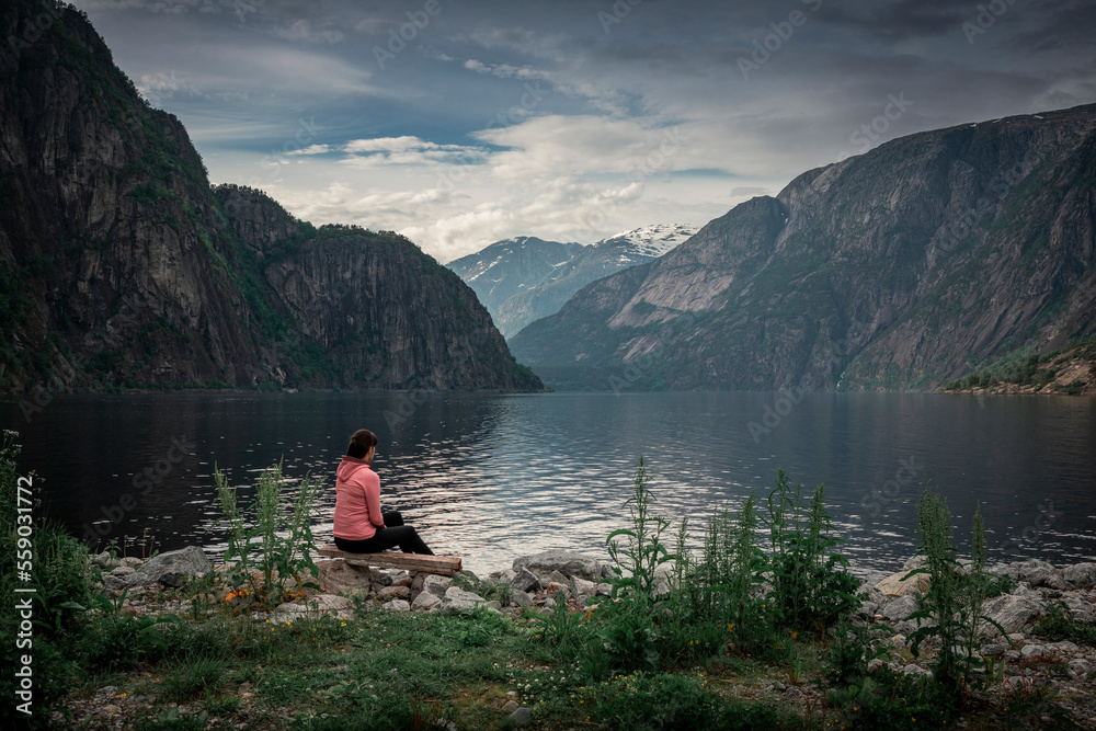 Woman sitting at waterfront of lake in the mountain landscape Eidfjord in Norway, looking into the fjord, clouds in the sky