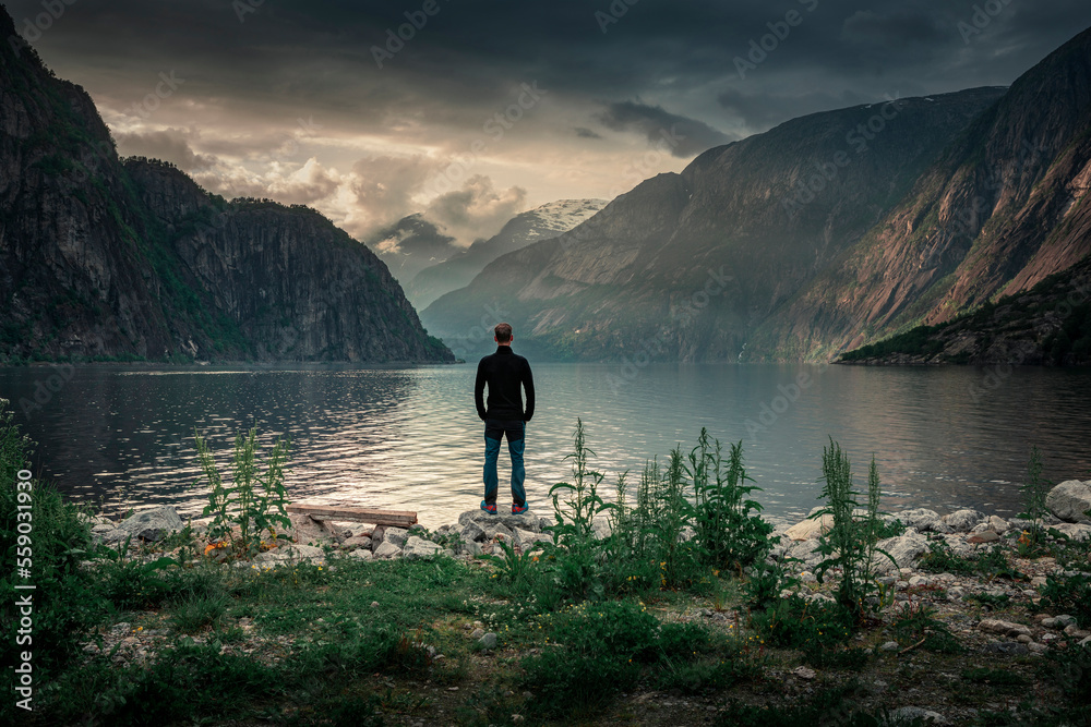 Man standing at waterfront of lake in the mountain landscape Eidfjord in Norway, looking into the fjord, clouds in the sky during sunset Man standing at waterfront of lake in the mountain landscape Eidfjord in Norway, looking into the fjord, clouds in the sky during sunset