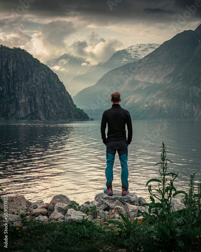 Man standing at waterfront of lake in the mountain landscape Eidfjord in Norway, looking into the fjord, clouds in the sky during sunset