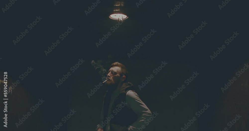 A young European man with beard in glasses smokes cigarette on dark background. SUPER slow motion 4k. Cigarette smoke close up. Gloomy tragic sad mood. glowing lamp hangs above person. Loft lift lamp A young European man with beard in glasses smokes cigarette on dark background. SUPER slow motion 4k. Cigarette smoke close up. Gloomy tragic sad mood. glowing lamp hangs above person. Loft lift lamp