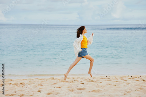 Sports woman runs along the beach in summer clothes on the sand in a yellow T-shirt and denim shorts white shirt flying hair ocean view