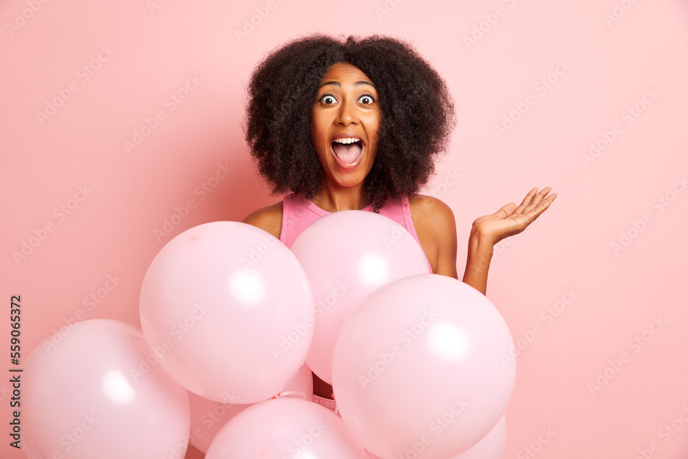 Overjoyed cute woman with curly hair screams loudly surrounded by helium balloons, raises palm and feels extremely happy, isolated over pink wall Overjoyed cute woman with curly hair screams loudly surrounded by helium balloons, raises palm and feels extremely happy, isolated over pink wall
