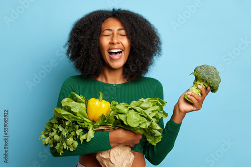 Indoor shot of curly haired young woman holds cluster of fresh vegetables and broccoli in a hand, laughs loudly, isolated over blue wall