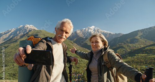 Mature caucasian couple travelling together, having a nordic walking hike in spring mountains, then stopping to take a picture on smartphone, spending time after retirement - recreational pursuit