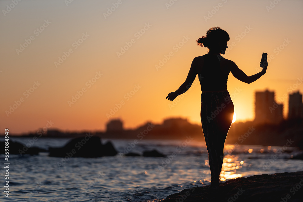woman making a self portrait at sunset on the beach, selfie with mobile phone, golden sunset, woman with mobile phone