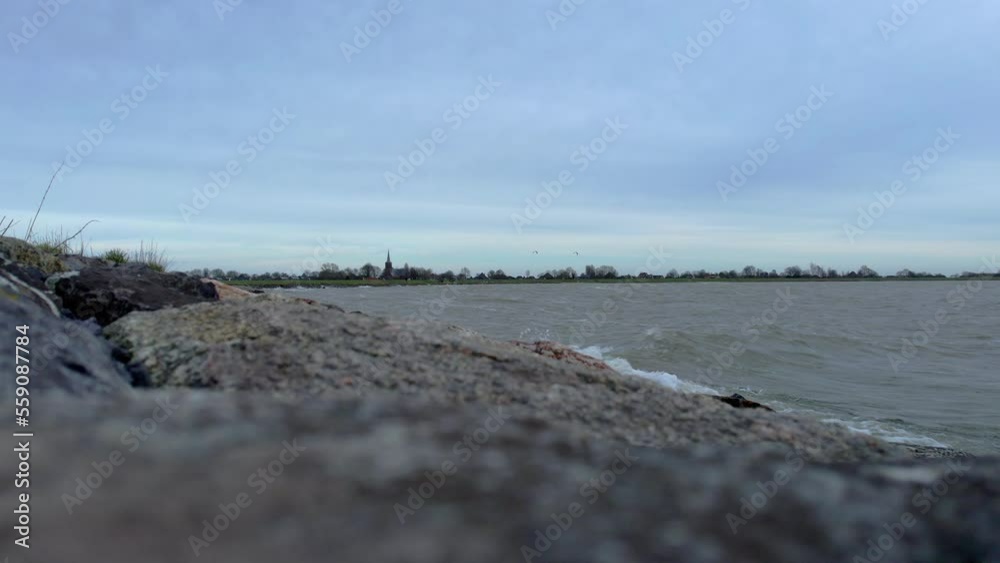 Static view at ground level over a lake with calm waves splashing on the boulders, grass blowing in the wind. In the background kitesurfers and a picturesque village with a church tower and trees Static view at ground level over a lake with calm waves splashing on the boulders, grass blowing in the wind. In the background kitesurfers and a picturesque village with a church tower and trees