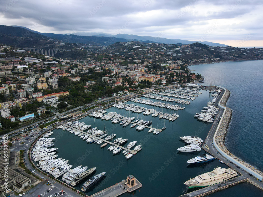 Fototapeta premium Aerial view of Sanremo, Italian city on the seashore in Liguria, north Italy. Drone flying along the port over beaches and boardwalk with palm trees and Birds Eye of yacht parking in San Remo, Italy.